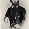 A black and white portrait photo of Mueller with faded edges. He has a full beard and is dressed in a three-piece suit, with medals hanging from his jacket. He has a book in his right hand and is holding a cutting from a plant in his left. He is looking past the camera into the distance.